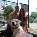 Smiling couple sitting with a white dog on a wooden deck outdoors, with a metal lattice fence and greenery in the background.