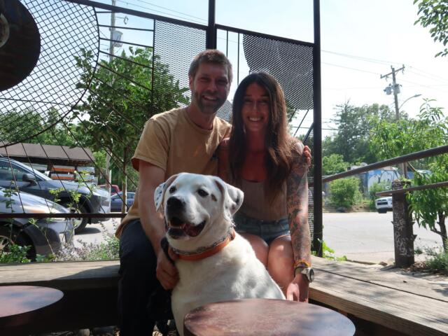 Smiling couple sitting with a white dog on a wooden deck outdoors, with a metal lattice fence and greenery in the background.