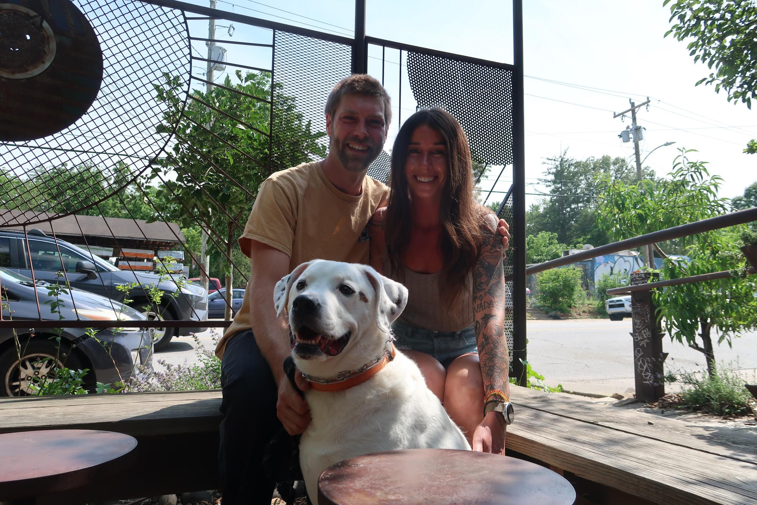 Smiling couple sitting with a white dog on a wooden deck outdoors, with a metal lattice fence and greenery in the background.