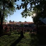 Outdoor festival with a crowd seated on grass under colorful bunting, trees providing shade and a bright blue sky.