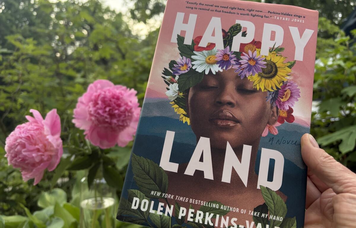 Close-up of a book cover showing a woman's face with a vibrant flower crown, outdoors among pink blooms; title reads 'HAPPY LAND'.