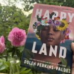 Close-up of a book cover showing a woman's face with a vibrant flower crown, outdoors among pink blooms; title reads 'HAPPY LAND'.