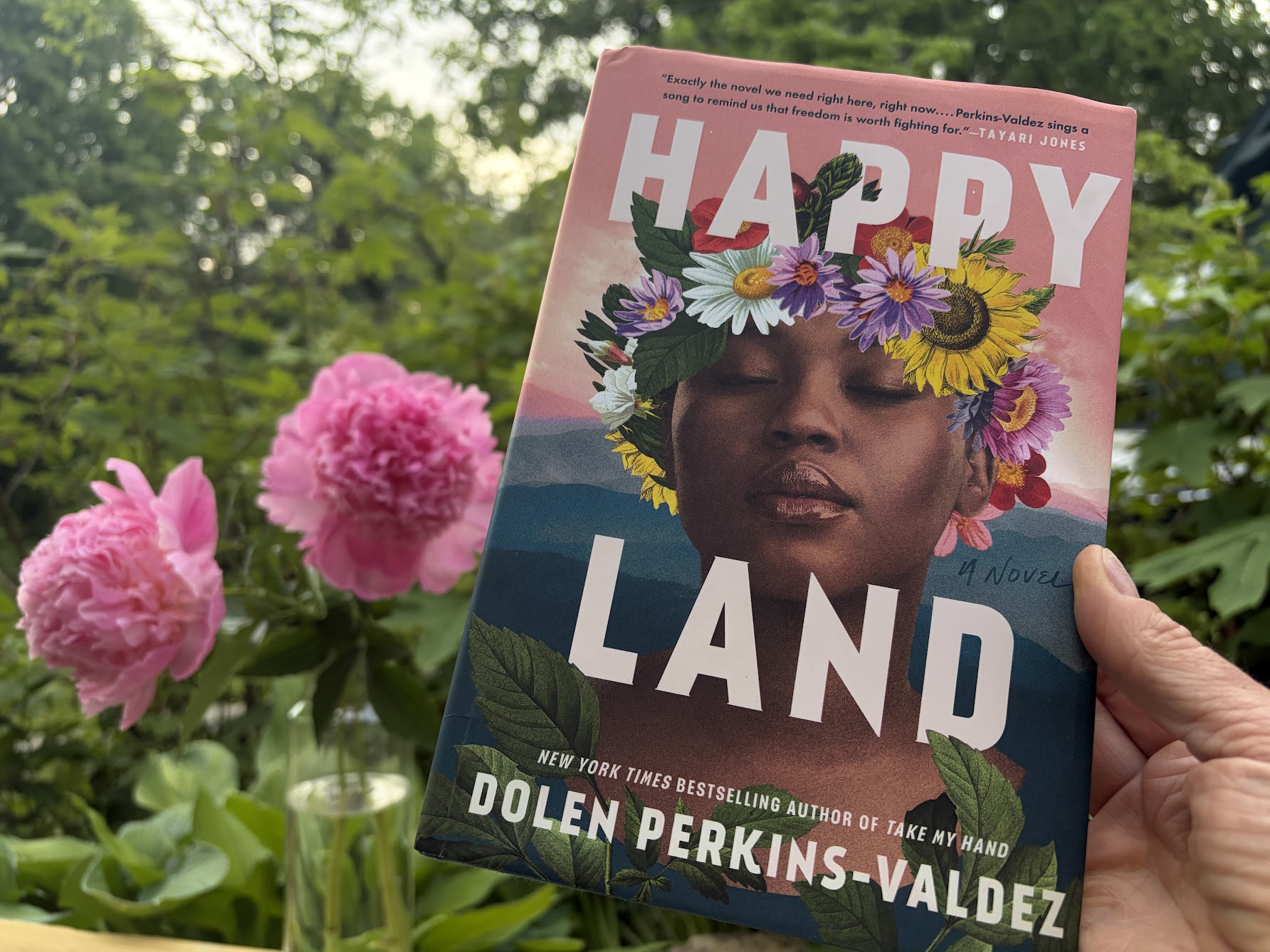Close-up of a book cover showing a woman's face with a vibrant flower crown, outdoors among pink blooms; title reads 'HAPPY LAND'.