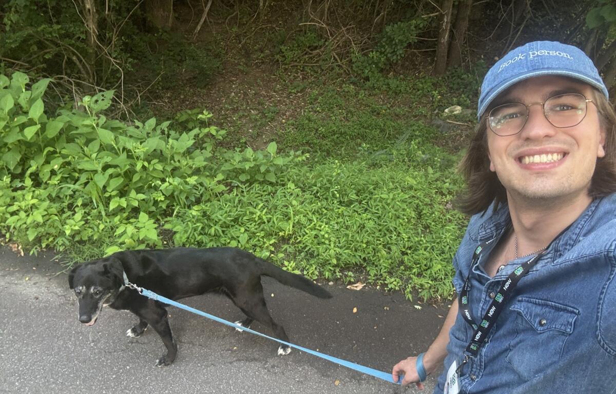 Person taking a selfie outdoors with a black dog on a blue leash; dense green shrubs behind them.