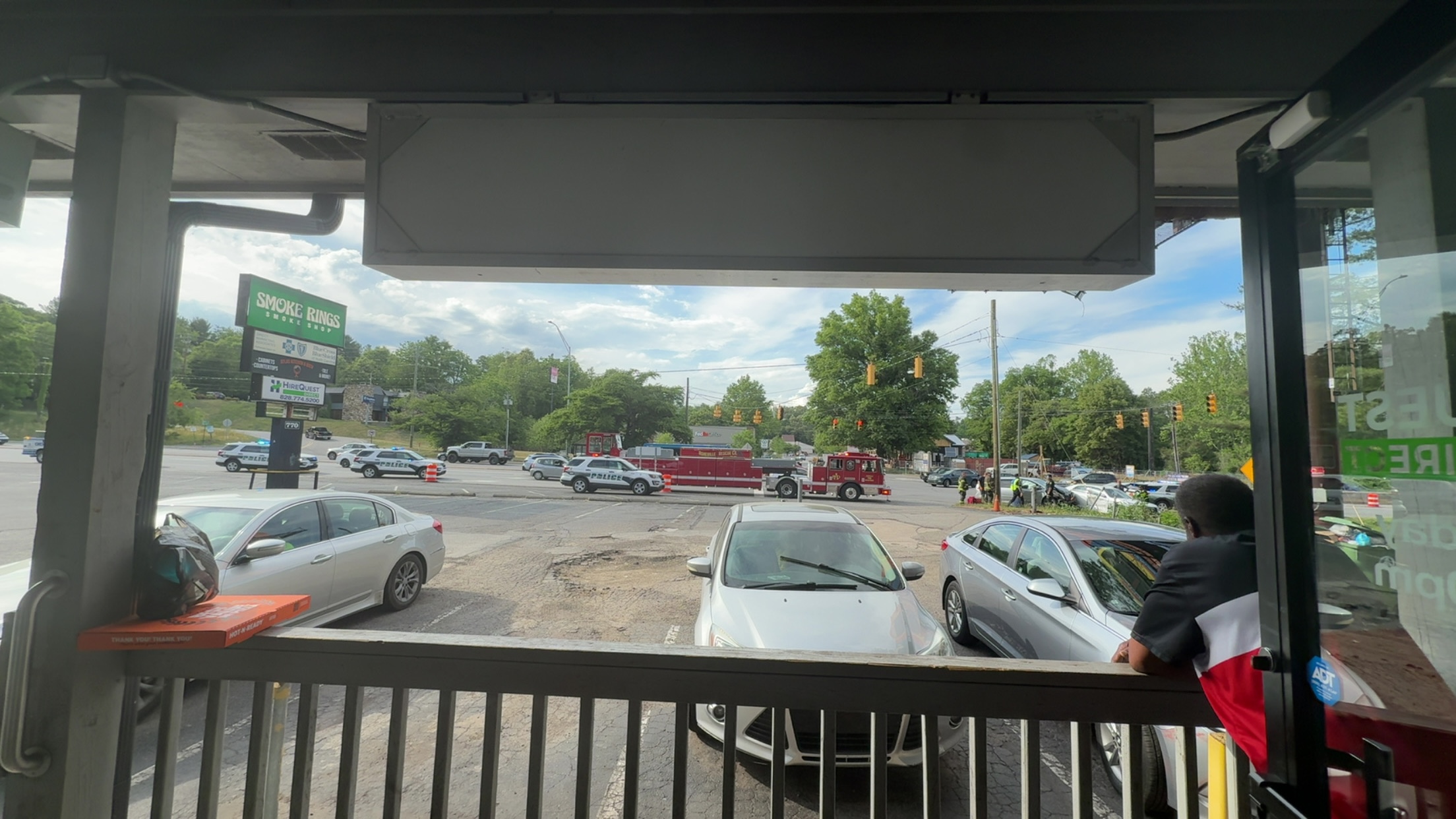 View from inside a storefront showing a busy intersection with a fire truck, police cars, and bystanders responding to an emergency.