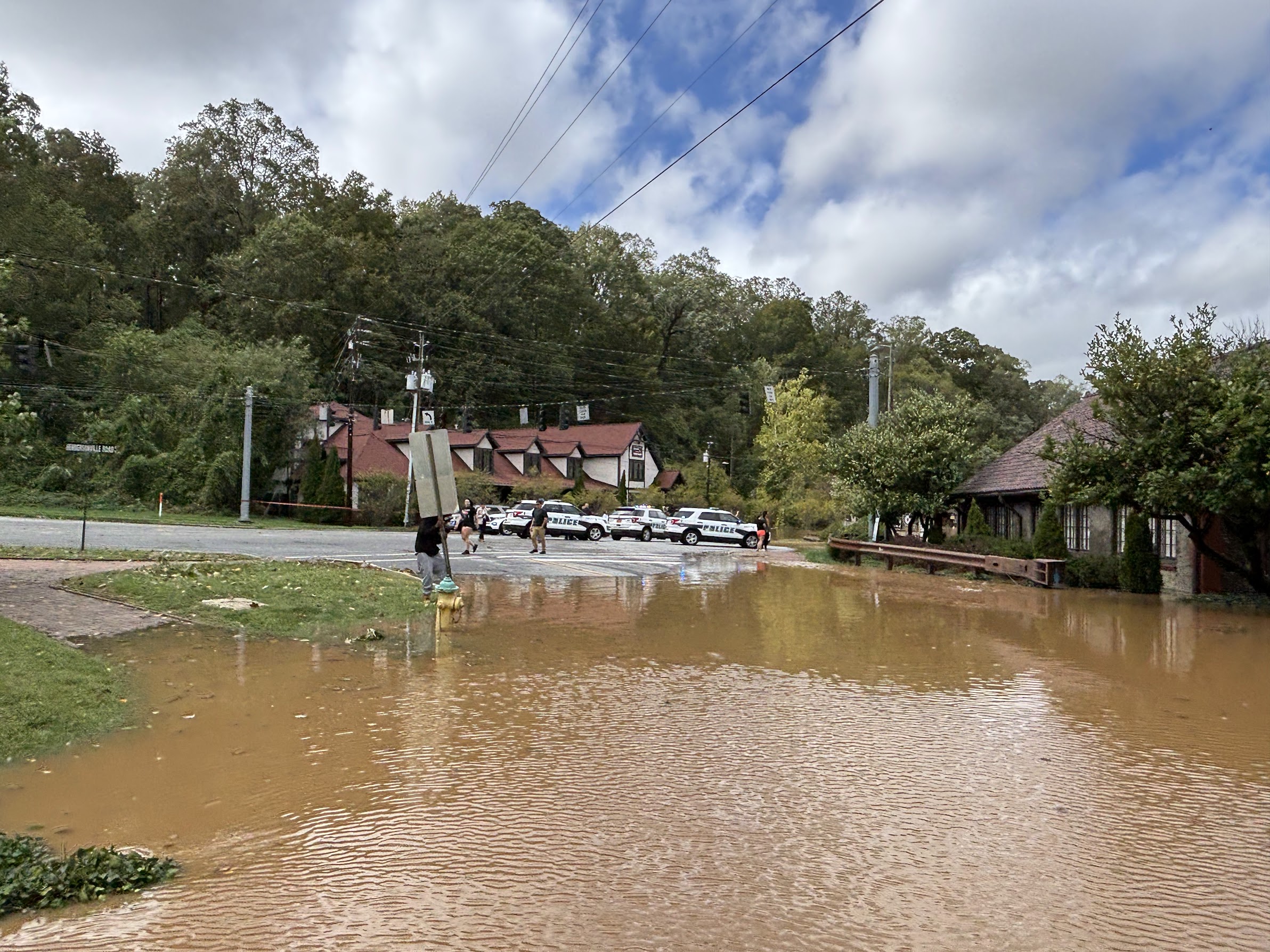 Flooded residential street with brown water, Police cars parked along the curb, and people standing nearby under a cloudy sky, power lines overhead.