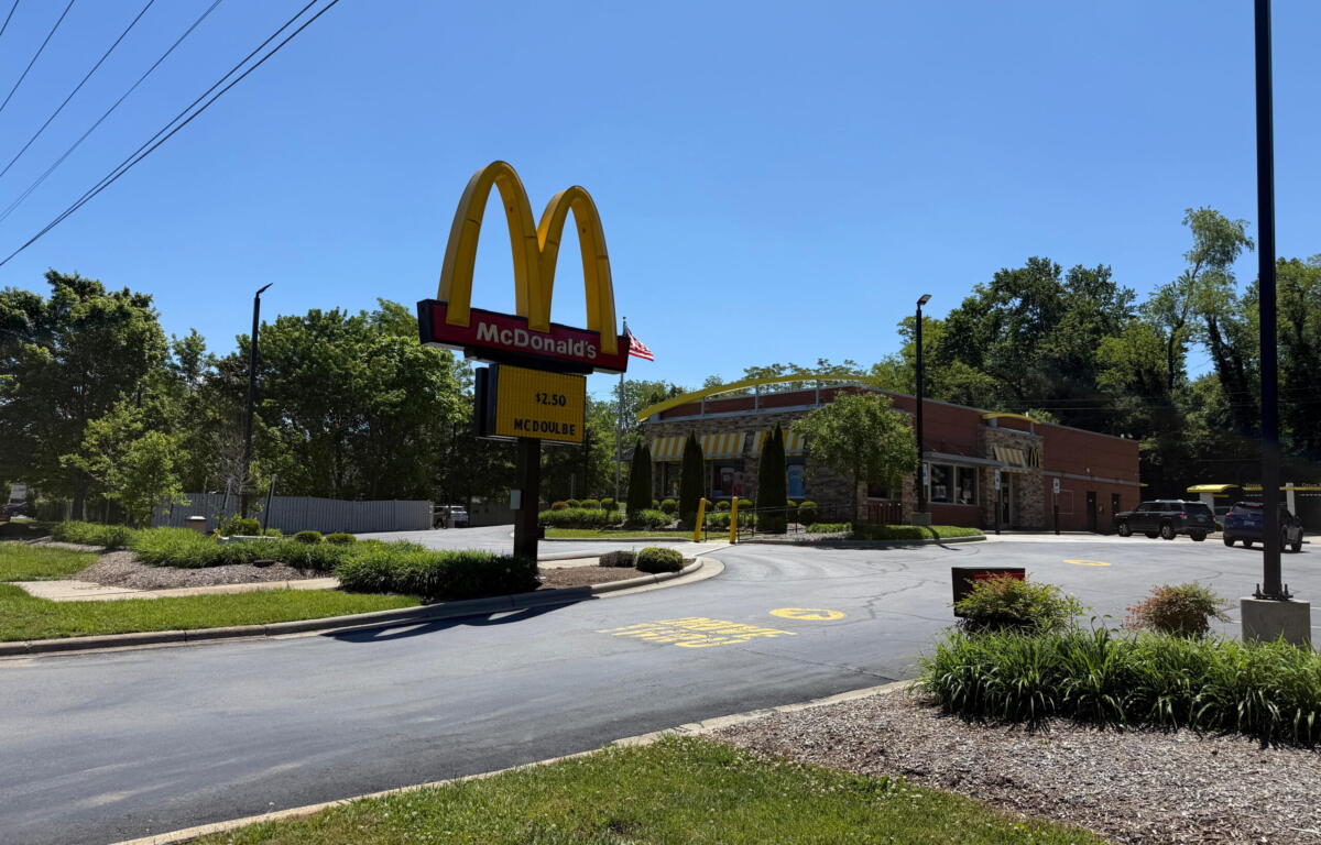 McDonald's sign with golden arches and red panel, restaurant building in background on a sunny day.