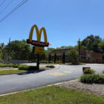 McDonald's sign with golden arches and red panel, restaurant building in background on a sunny day.