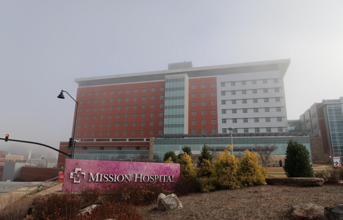Exterior view of Mission Hospital campus: a multi-story building with red brick and white panels, glass atrium, seen from the street.