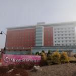 Exterior view of Mission Hospital campus: a multi-story building with red brick and white panels, glass atrium, seen from the street.