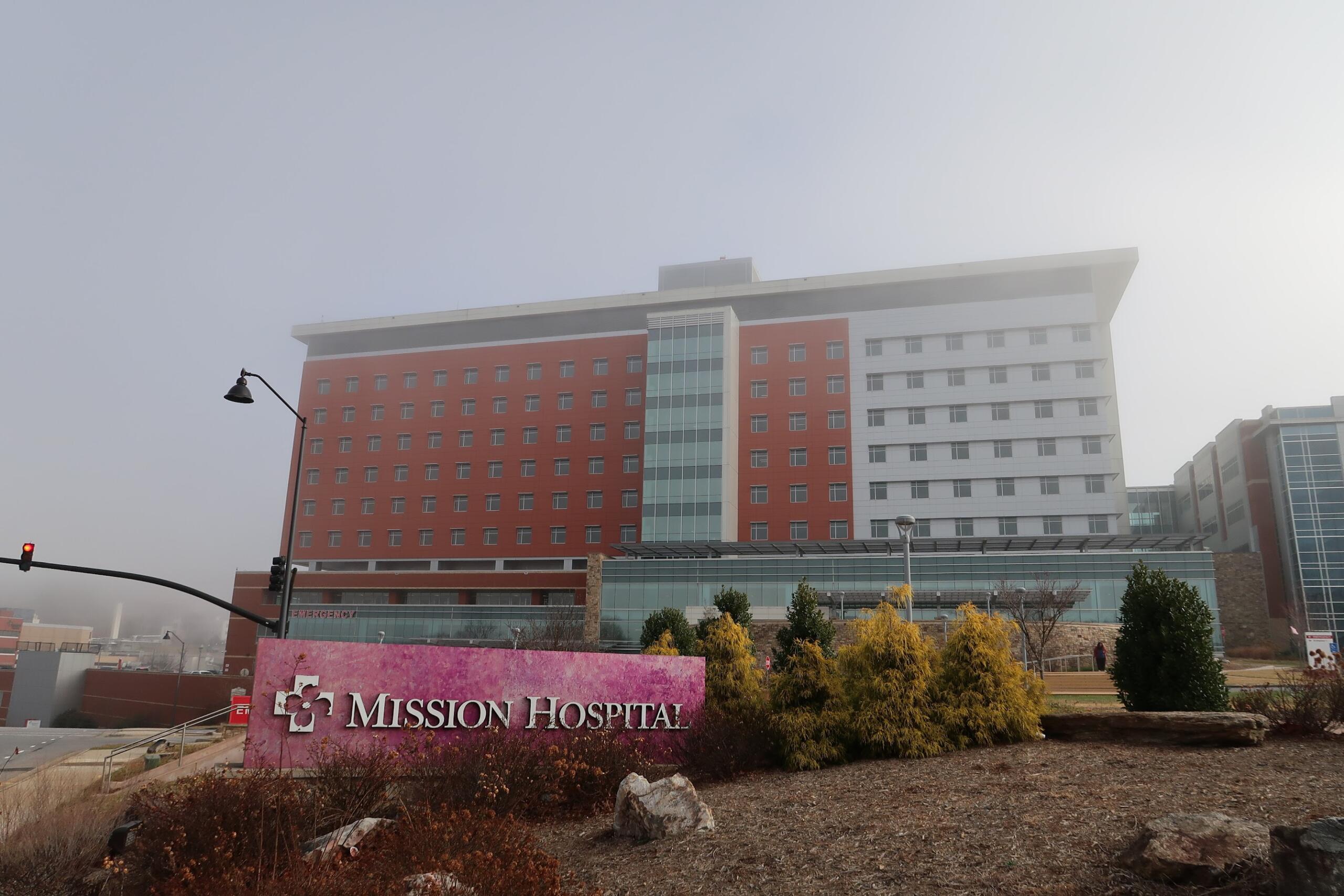 Exterior view of Mission Hospital campus: a multi-story building with red brick and white panels, glass atrium, seen from the street.