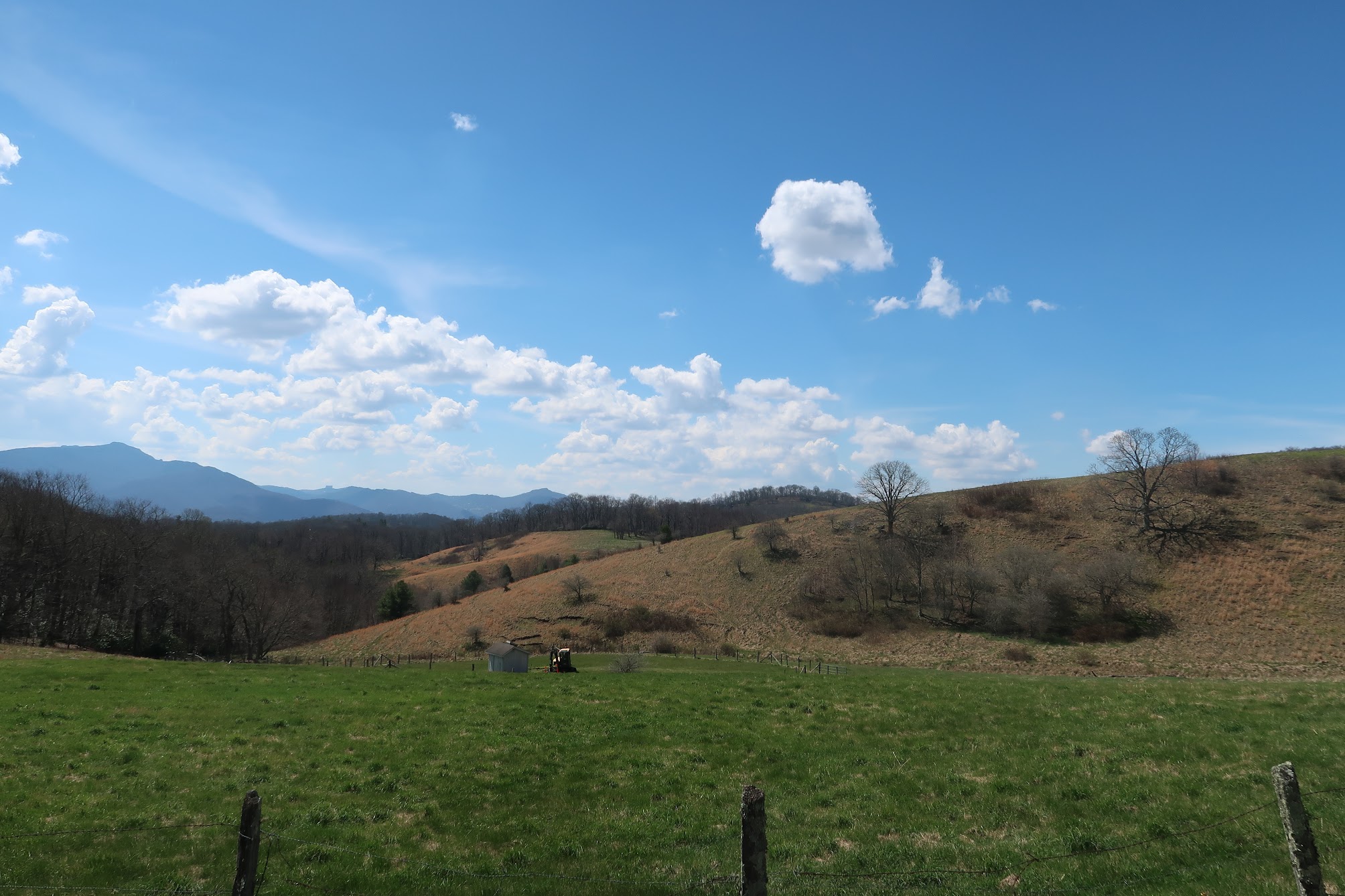 Open rural landscape with rolling brown hills, leafless trees, a wooden fence in the foreground, and a bright blue sky with scattered clouds.