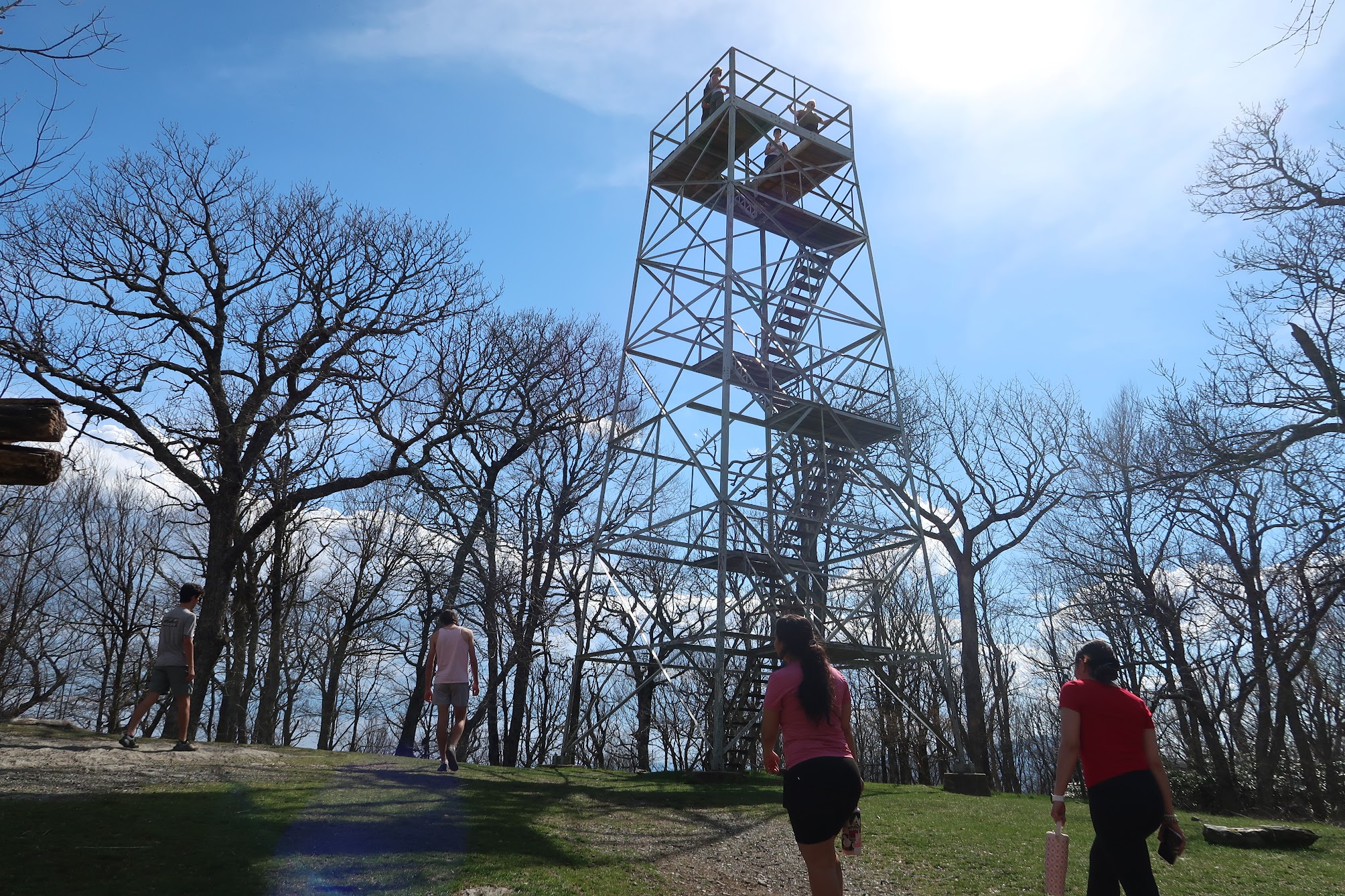 Runners on a dirt path beside a tall metal observation tower in a leafless forest on a sunny day in springtime light.