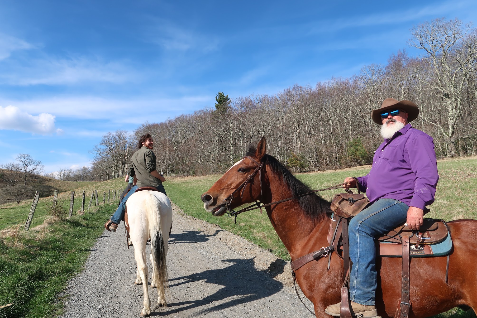 Two riders on horses along a rural gravel path, with leafless trees and a bright blue sky.