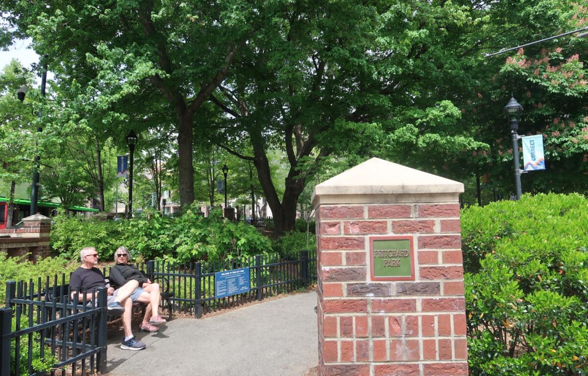 Two people sit on a black metal bench beside a brick column with a Pritchard Park plaque, in a green, leafy city park with lampposts and banners in the background.