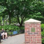 Two people sit on a black metal bench beside a brick column with a Pritchard Park plaque, in a green, leafy city park with lampposts and banners in the background.