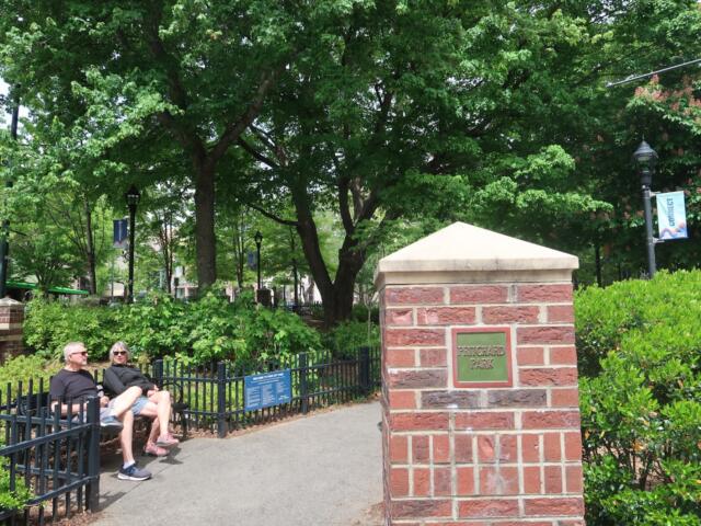 Two people sit on a black metal bench beside a brick column with a Pritchard Park plaque, in a green, leafy city park with lampposts and banners in the background.