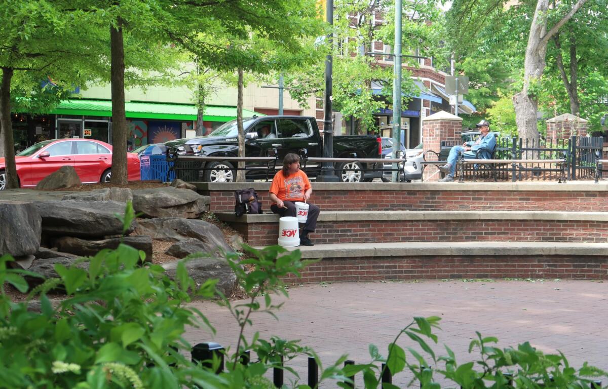 Man in an orange shirt sits on brick steps of a park, with two white buckets in front and greenery behind.