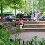 Man in an orange shirt sits on brick steps of a park, with two white buckets in front and greenery behind.