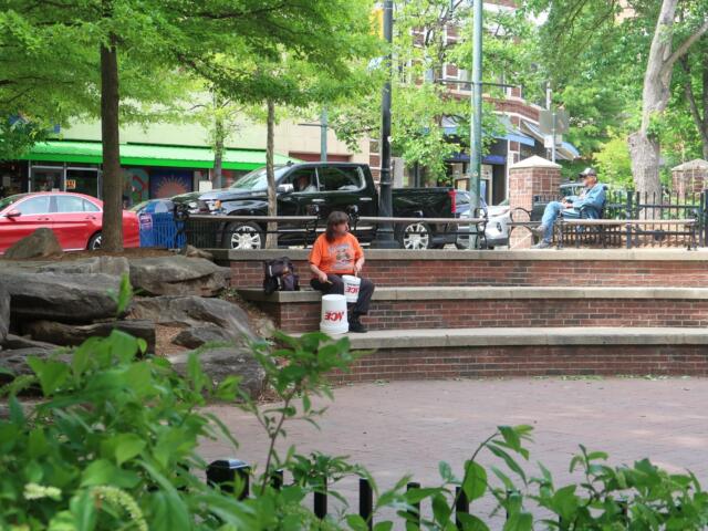 Man in an orange shirt sits on brick steps of a park, with two white buckets in front and greenery behind.