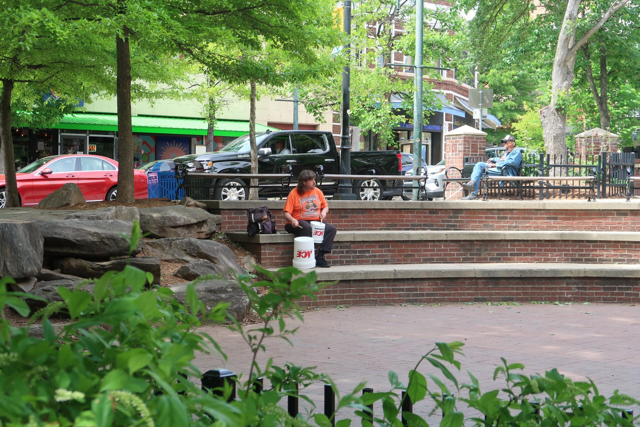 Man in an orange shirt sits on brick steps of a park, with two white buckets in front and greenery behind.