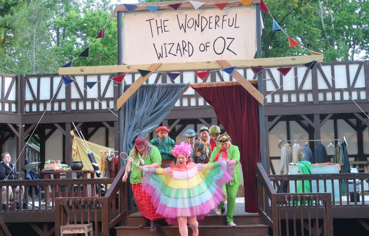 Colorful performers on a wooden outdoor stage under a banner reading 'The Wonderful Wizard of Oz' in front of a crowd.