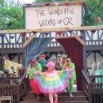 Colorful performers on a wooden outdoor stage under a banner reading 'The Wonderful Wizard of Oz' in front of a crowd.