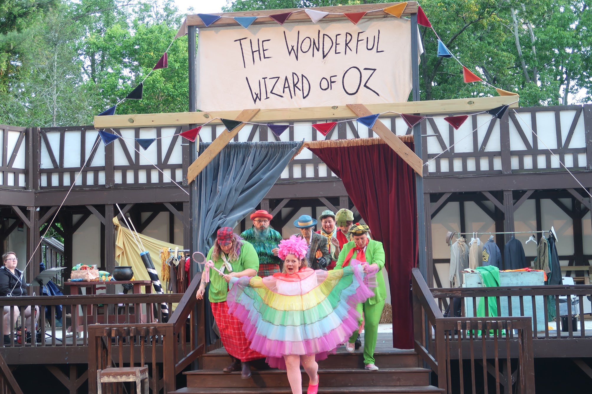 Colorful performers on a wooden outdoor stage under a banner reading 'The Wonderful Wizard of Oz' in front of a crowd.
