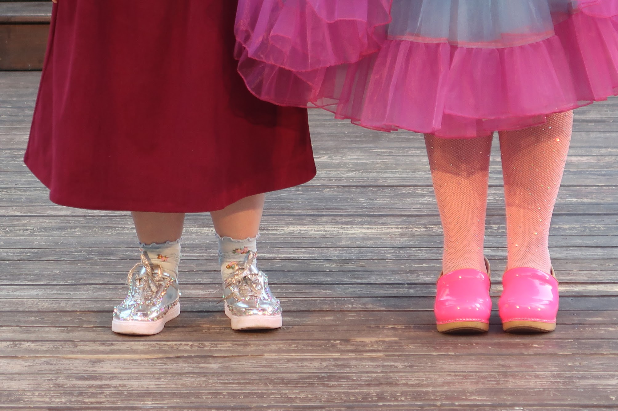 Two children stand on a wooden deck in festive outfits: one in a burgundy dress with glittery silver boots, the other in a pink tutu and bright pink shoes.