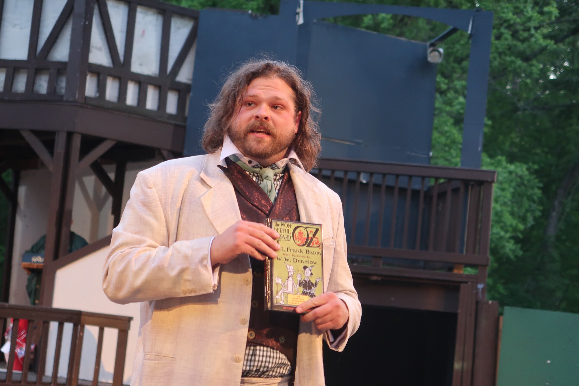 Man with long hair and beard in a light beige suit holding a vintage illustrated book on an outdoor stage.