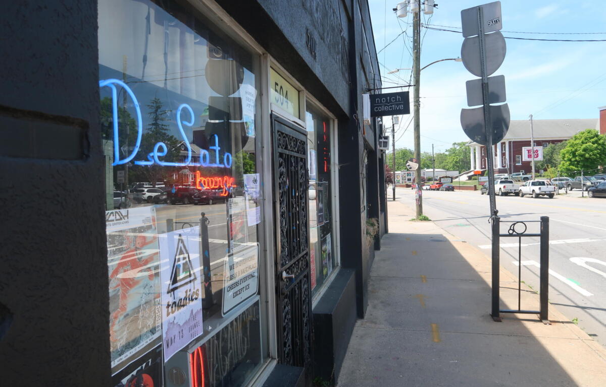 Storefront window display with a bright blue neon 'Detroit' sign and posters, on a sunny street façade.