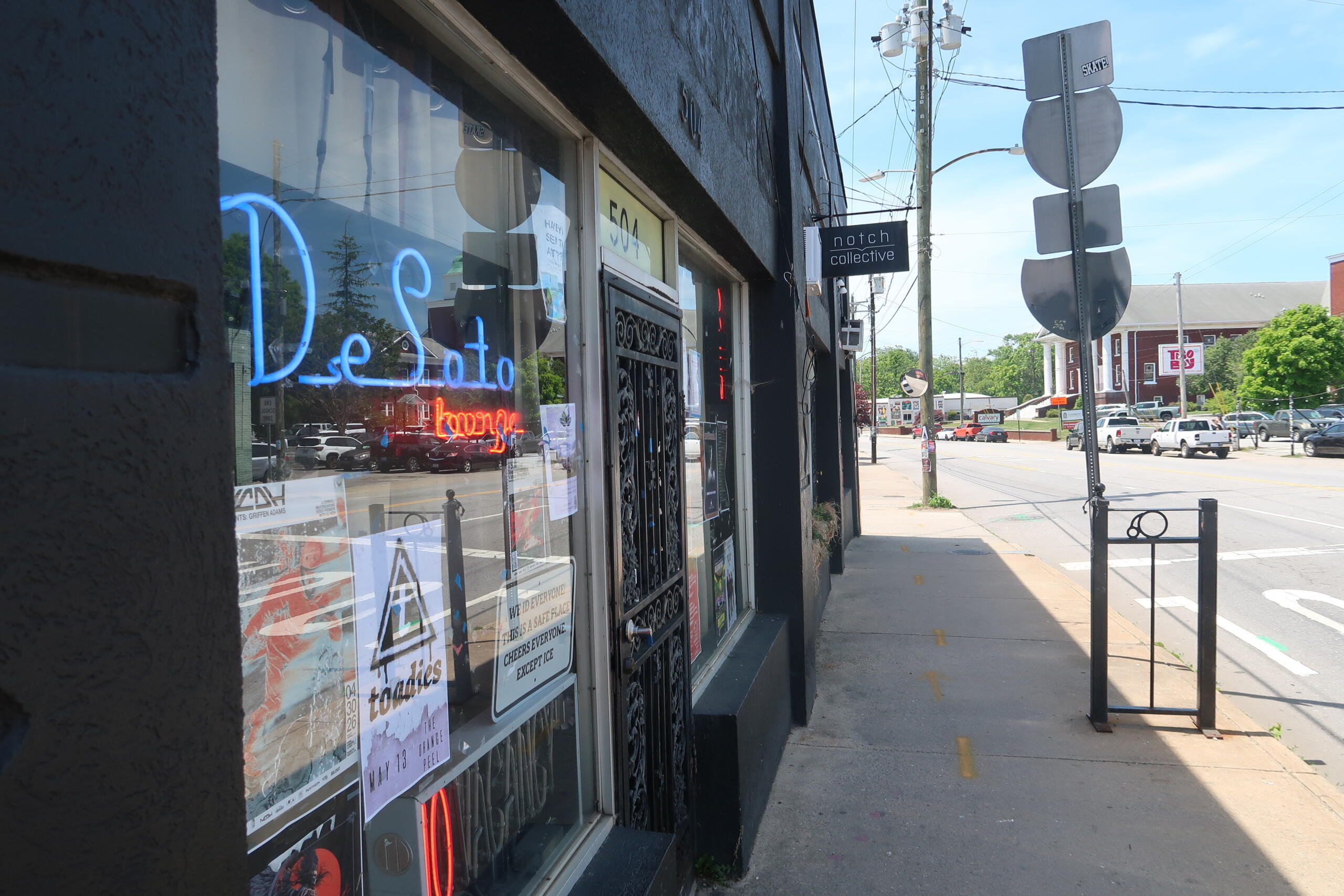 Storefront window display with a bright blue neon 'Detroit' sign and posters, on a sunny street façade.