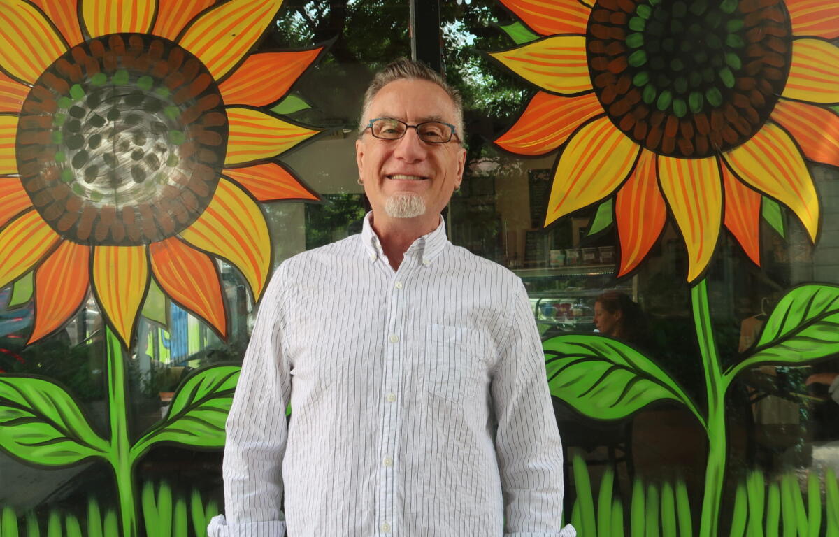 Smiling man with glasses stands in front of a bright mural of oversized orange-yellow sunflowers with green leaves.