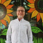 Smiling man with glasses stands in front of a bright mural of oversized orange-yellow sunflowers with green leaves.