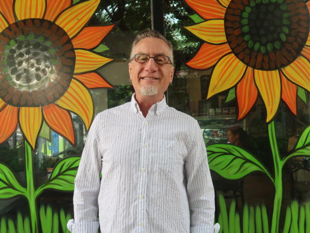 Smiling man with glasses stands in front of a bright mural of oversized orange-yellow sunflowers with green leaves.