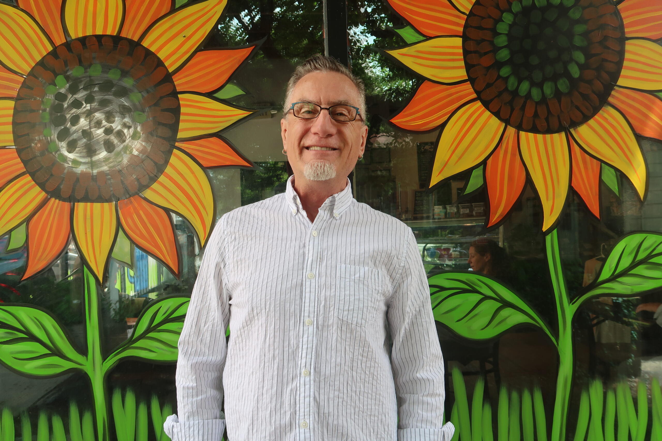 Smiling man with glasses stands in front of a bright mural of oversized orange-yellow sunflowers with green leaves.