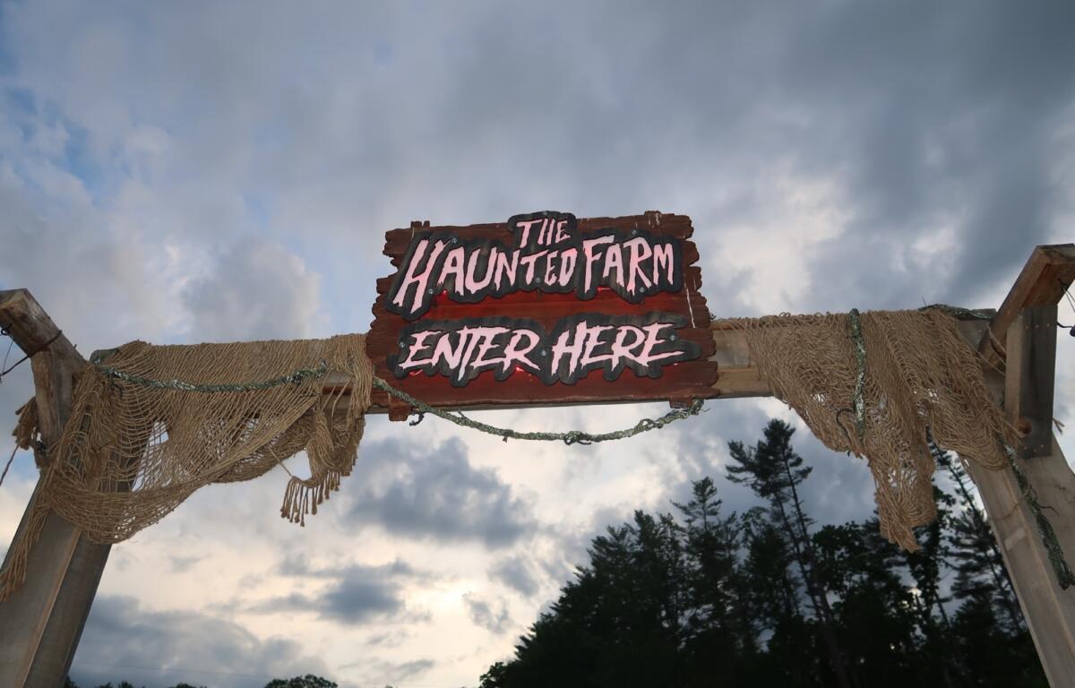 Wooden sign reading 'The Haunted Farm — Enter Here' hanging over rope netting against a cloudy sky.