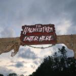 Wooden sign reading 'The Haunted Farm — Enter Here' hanging over rope netting against a cloudy sky.