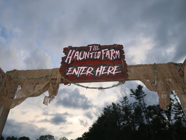 Wooden sign reading 'The Haunted Farm — Enter Here' hanging over rope netting against a cloudy sky.