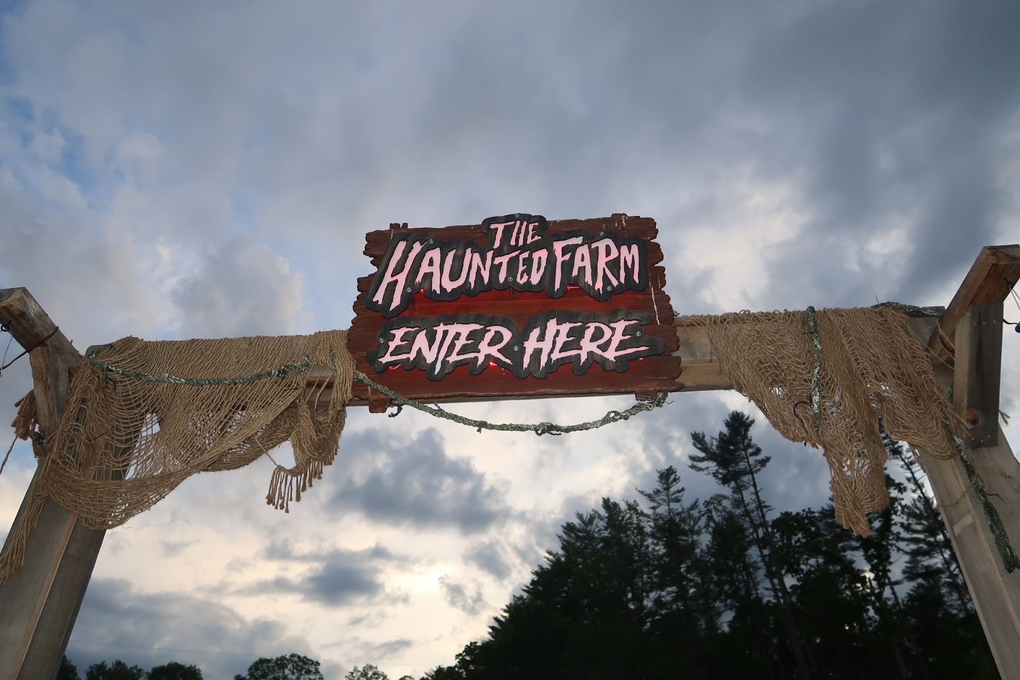 Wooden sign reading 'The Haunted Farm — Enter Here' hanging over rope netting against a cloudy sky.