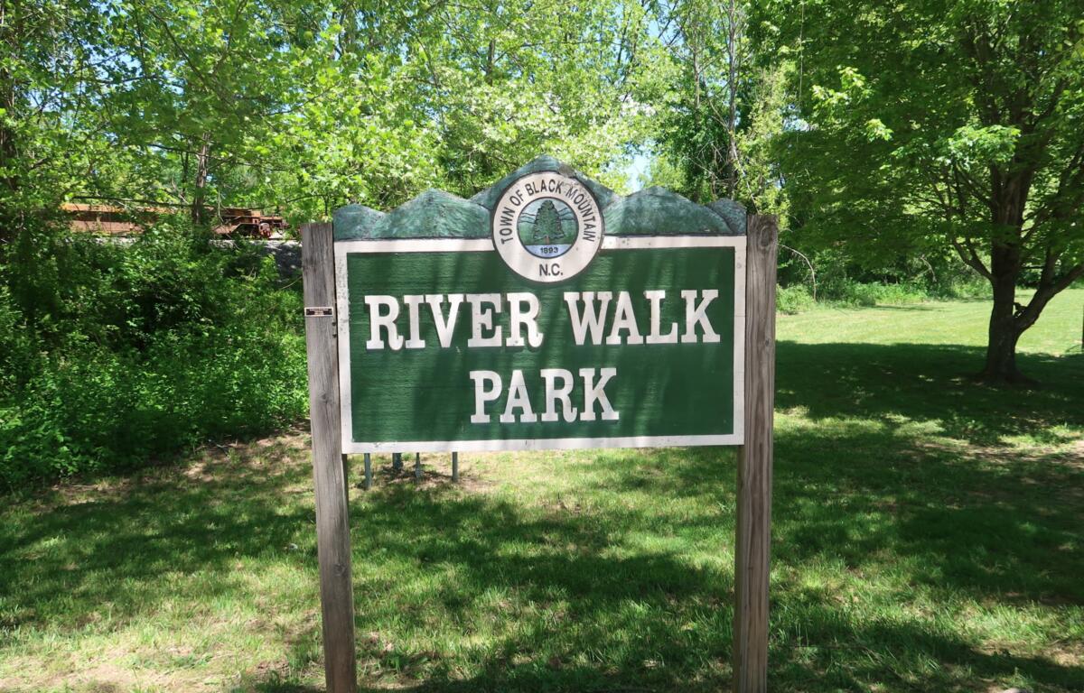 Sign for River Walk Park in a green wooded area under sunny sky, with trees surrounding.