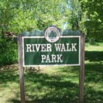 Sign for River Walk Park in a green wooded area under sunny sky, with trees surrounding.