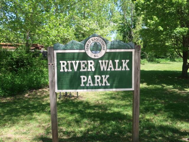 Sign for River Walk Park in a green wooded area under sunny sky, with trees surrounding.