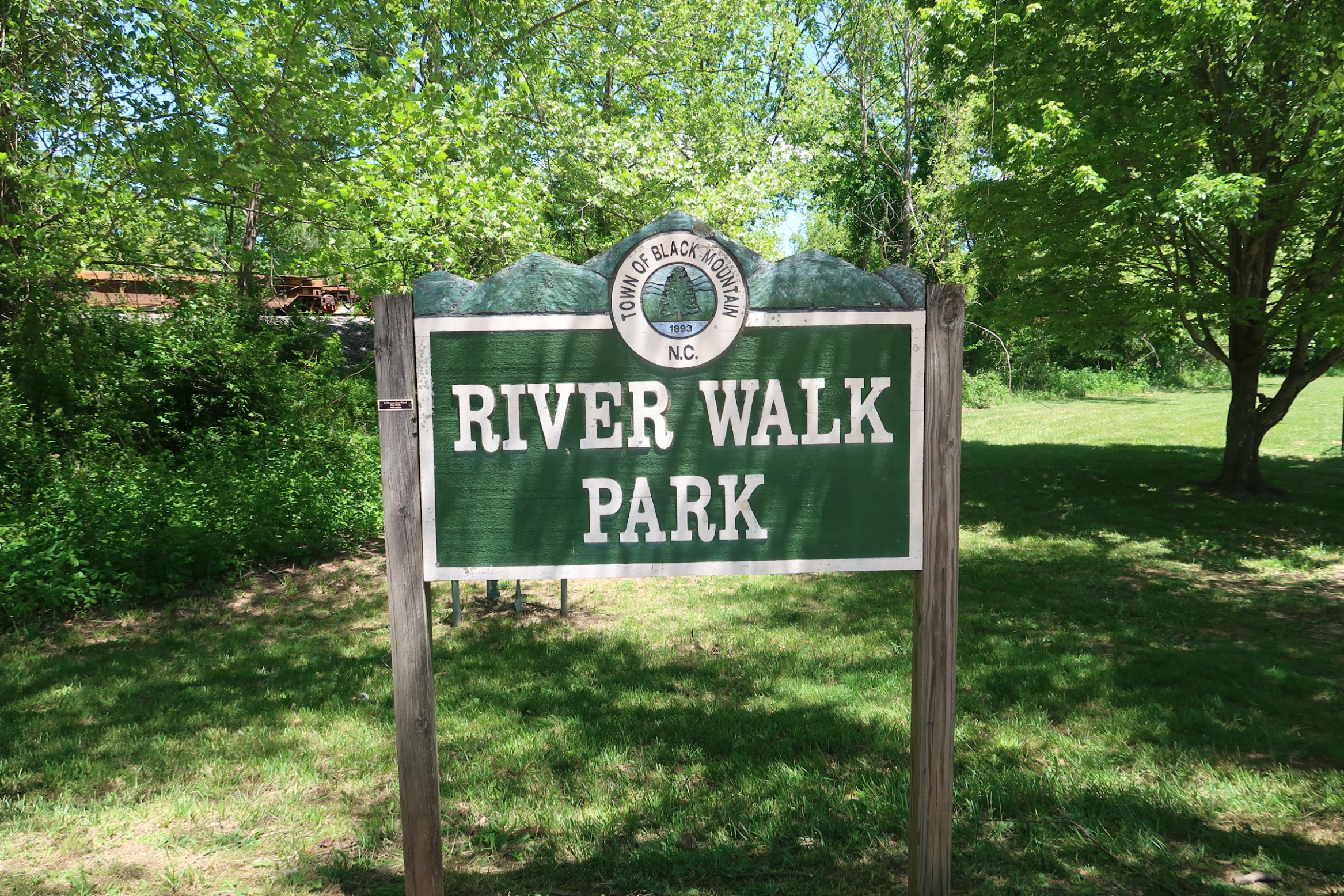 Sign for River Walk Park in a green wooded area under sunny sky, with trees surrounding.