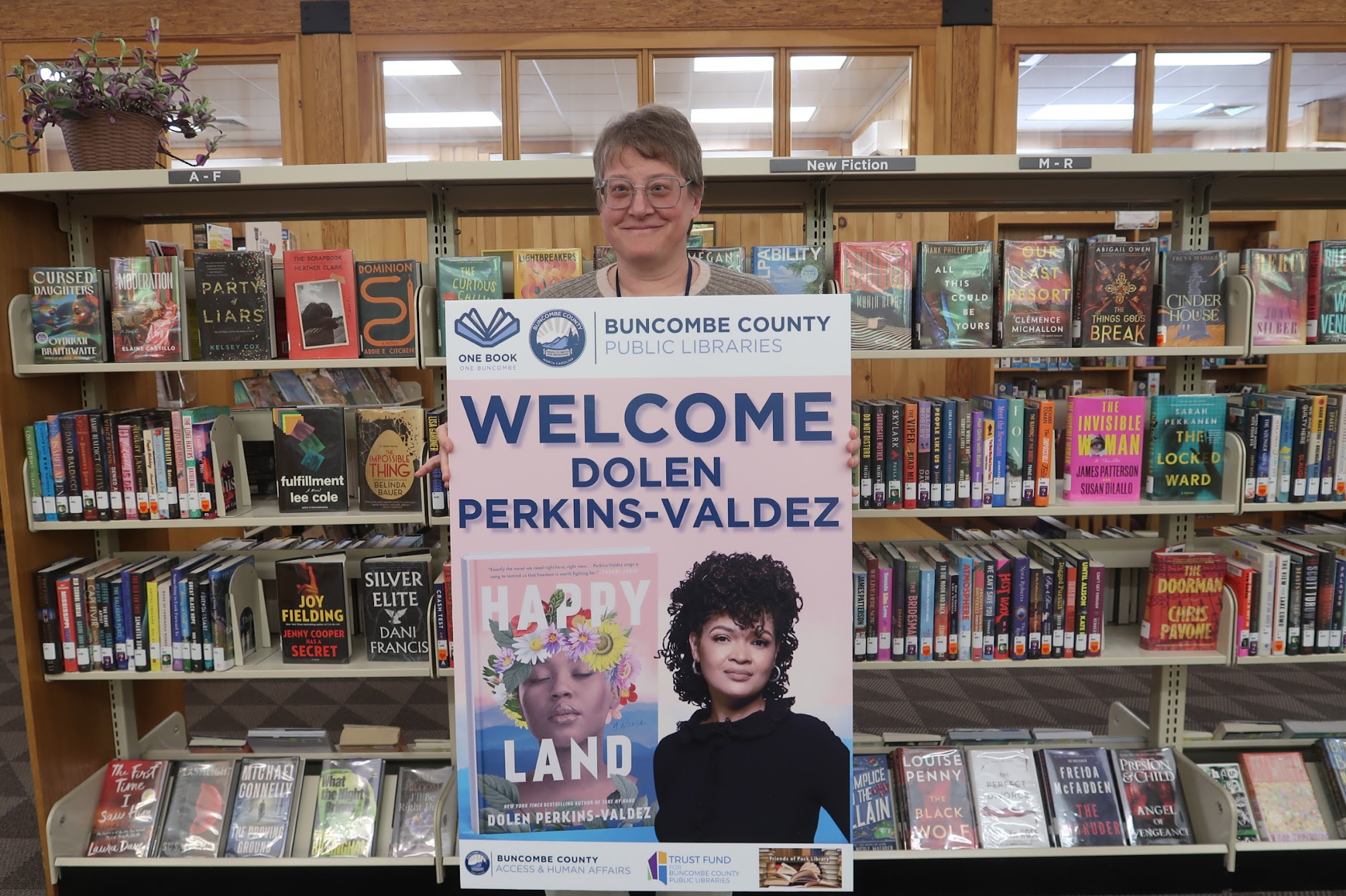 Person with short gray hair and glasses stands behind a large 'Welcome' poster in a library, surrounded by shelves of books.