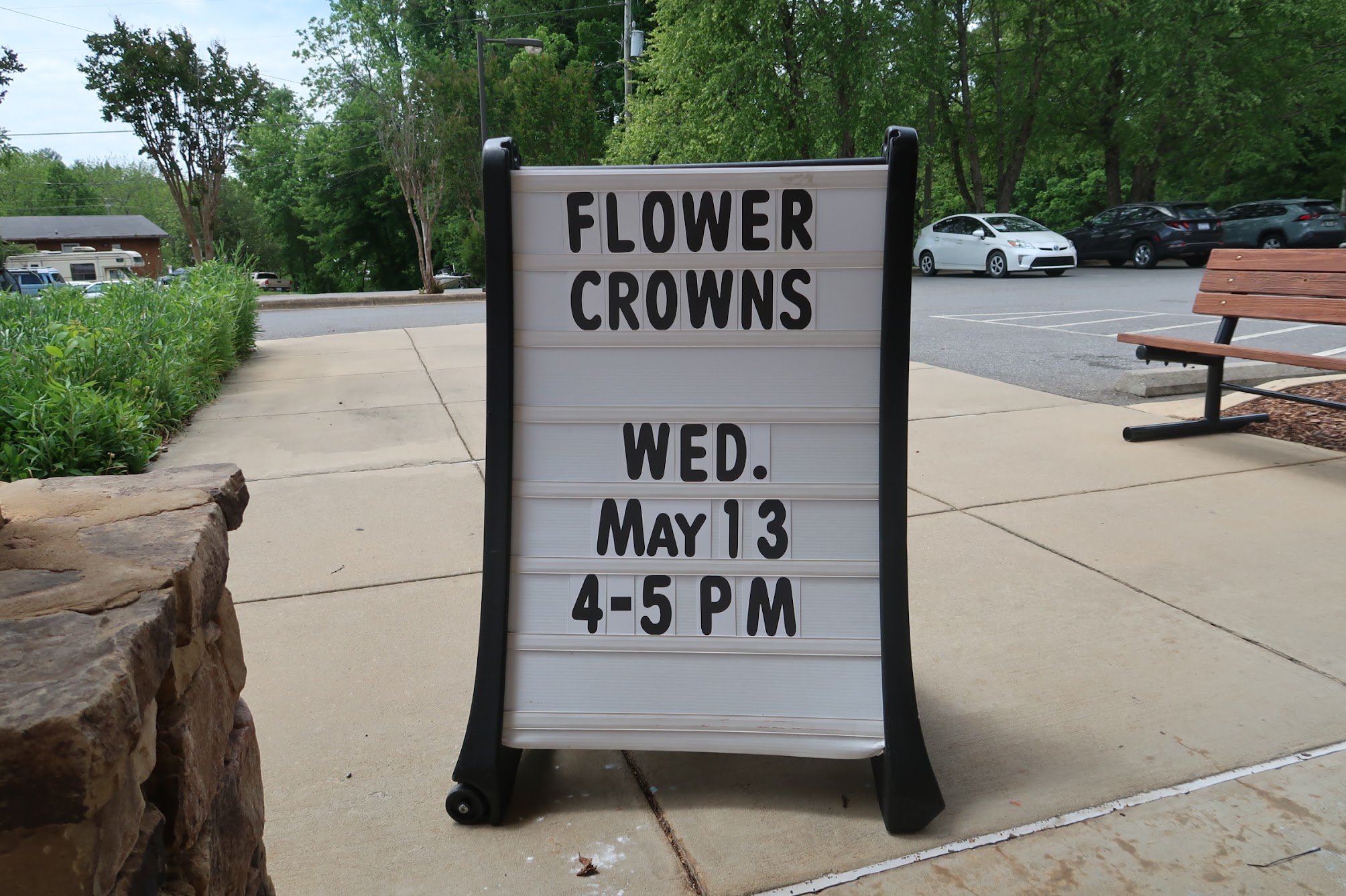 Outdoor roadside sign advertising a 'FLOWER CROWNS' event, with date and time listed (Wed May 13, 4–5 PM).