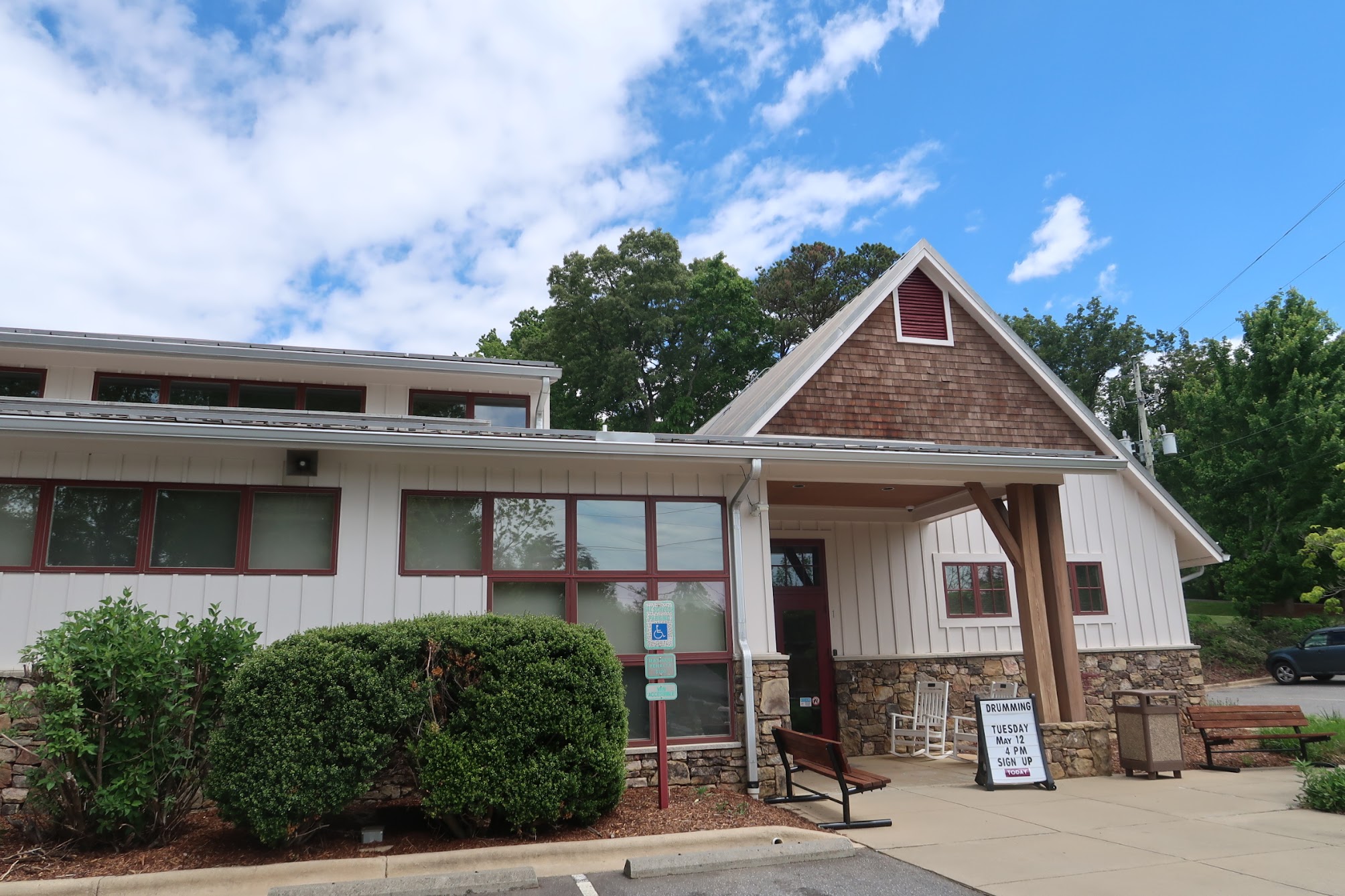 Front of a light-siding building with red-trimmed windows, a brown shingle gable, and outdoor seating under a covered area; a drumming event sign near the entrance.