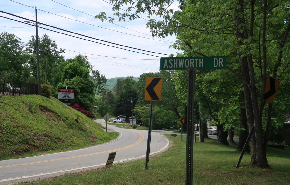 Curved road lined with trees and power lines; Ashworth Dr street sign visible with yellow chevron signs nearby.