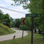Curved road lined with trees and power lines; Ashworth Dr street sign visible with yellow chevron signs nearby.