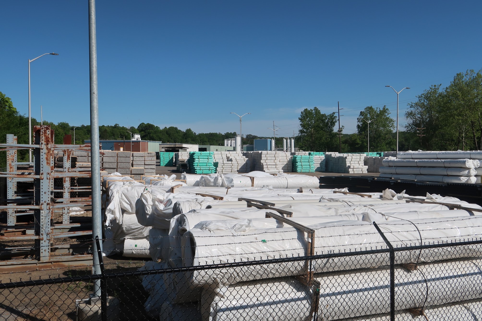 Open storage yard with stacked wrapped bundles, pallets, and rusted metal framework under a clear blue sky, fenced in